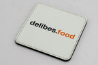 Posavasos cuadrado blanco con logotipo delibes.Food negro y naranja.