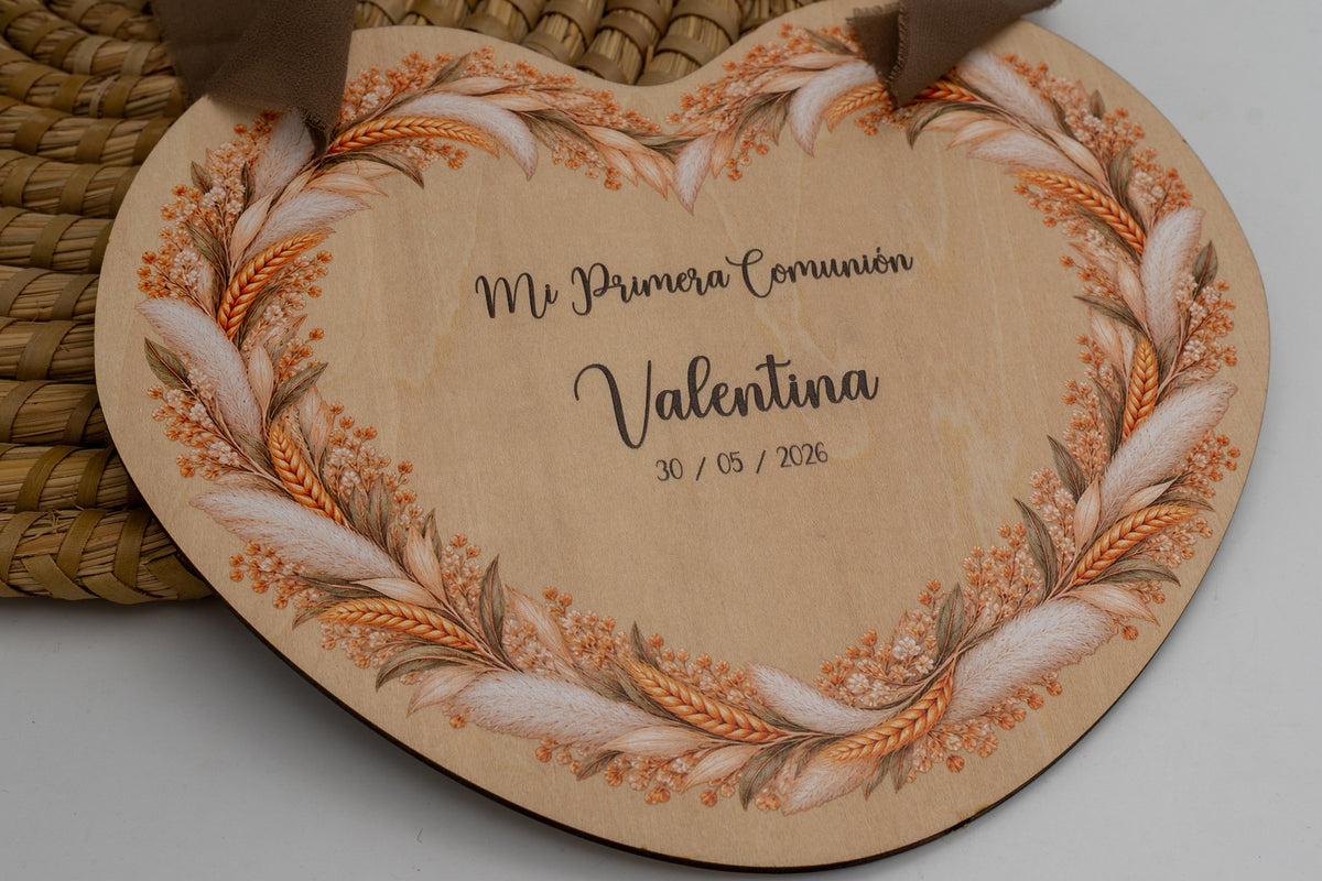 Cartel de comuñón de madeira en forma de corazón con coroa de espigas e herba da pampa, co texto «Mi Primera Comunión», o nome de exemplo Valentina e a data, colgado cunha cinta marrón.
