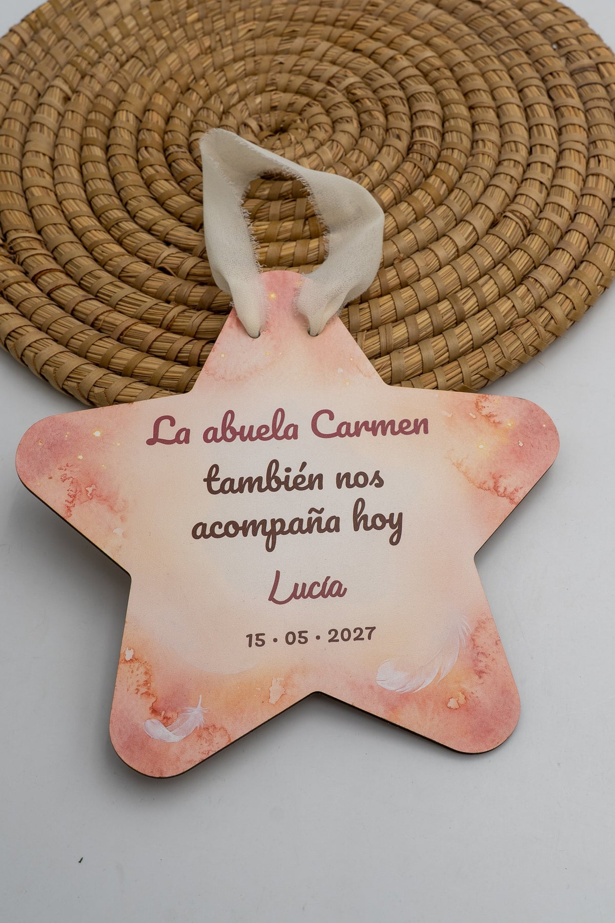 Estrela de comuñón con fondo de acuarela rosa e plumas brancas, a frase «La abuela Carmen también nos acompaña hoy», o nome de exemplo Lucía e a data, con cinta crema.