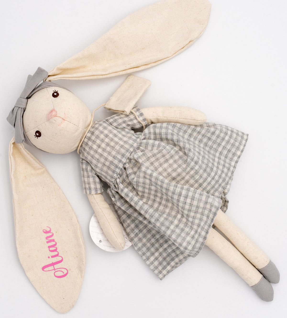 Muñeca conejita gris tumbada, con el nombre Ariane en la oreja y el vestido de cuadros visible.
