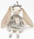 Muñeca de trapo conejita gris sentada, con el nombre Eugenia en una oreja, vestido de cuadros y bolso de tela.