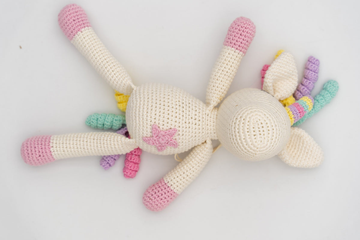 Vista cenital del unicornio de crochet crudo con patas rosas, cuerno y melena multicolor.