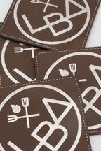 Detalle de posavasos cadrados marróns con logotipo branco e costuras.