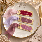 Tres marcasitios de metacrilato espejo dorado en granate, malva y rosa lavanda, con cintas chiffon coordinadas, sobre plato Picasita.