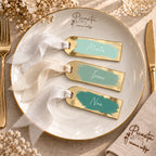 Tres marcasitios de metacrilato espejo dorado en verde agua, verde salvia y verde eucalipto, con cintas chiffon coordinadas, sobre plato Picasita.